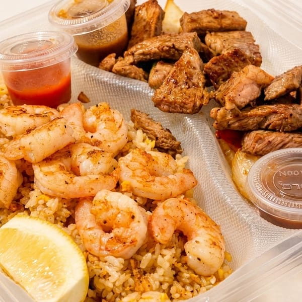 Shrimp fried rice with a lemon wedge and steak with onions, served in a clear to-go container.