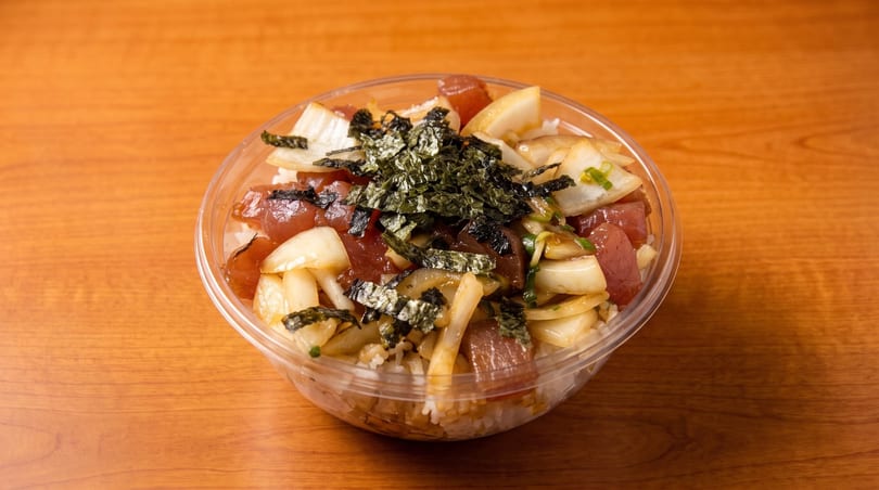 A clear plastic bowl of poke with tuna, onions, and seaweed flakes on a wooden table.