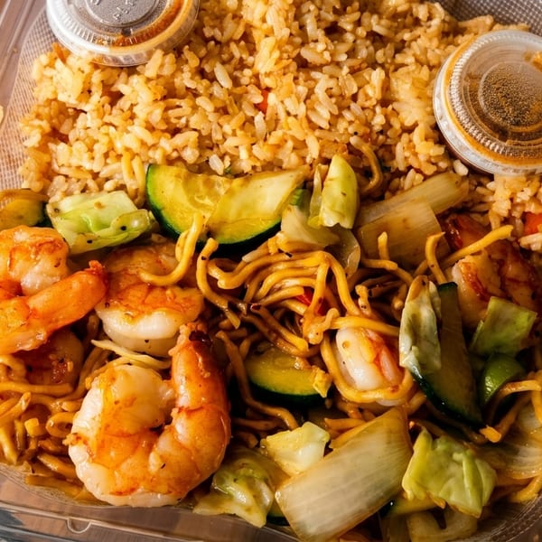 Shrimp, noodles, and vegetables with fried rice and sauce in a clear takeout container.