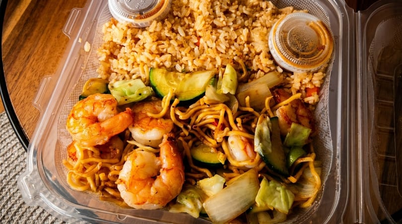 Shrimp, noodles, and vegetables with fried rice and two sauce containers in a plastic takeout box.