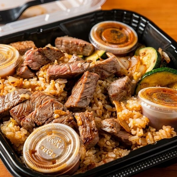 Takeout container filled with steak, fried rice, zucchini, and three sauce cups on a wooden table.