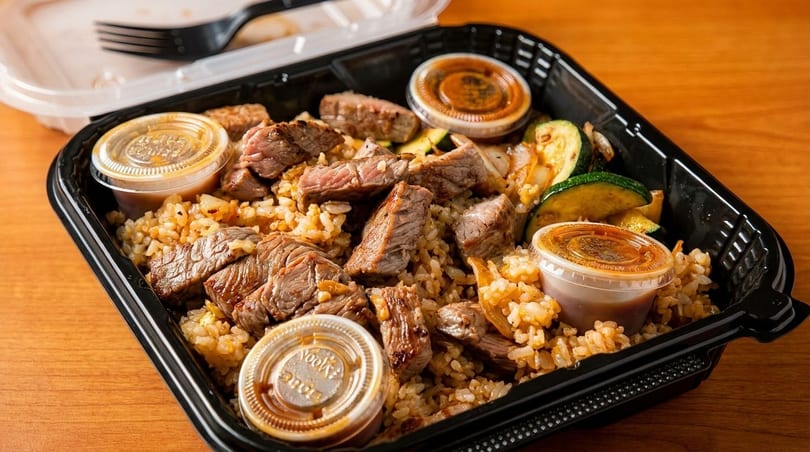 Steak and rice with zucchini and three sauce containers in a black takeout box.