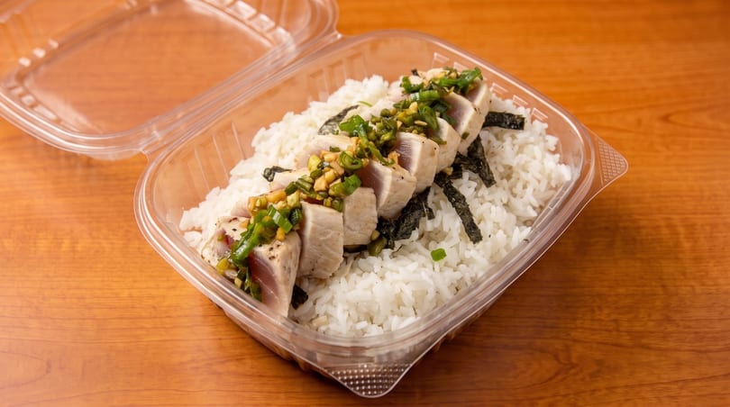 Seared tuna with green onions and garlic over white rice and seaweed in a plastic takeout container.