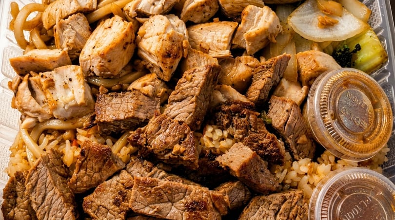 Close-up of a takeout container filled with grilled chicken, steak, noodles, rice, and vegetables.