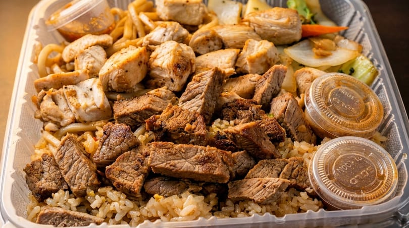 Close-up of a takeout container filled with steak, chicken, noodles, rice, and two sauce cups.