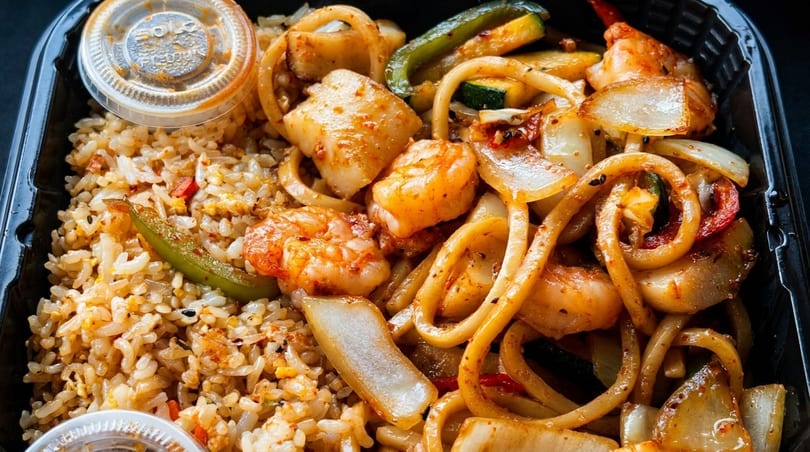 Hibachi shrimp, scallops, and noodles with fried rice and vegetables in a black takeout container.